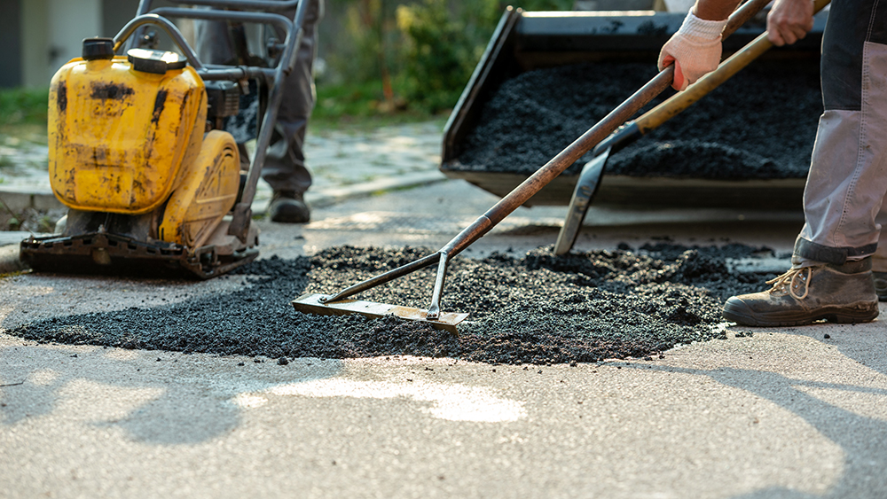 Two workers repairing damaged asphalt using a vibratory plate compactor and rake, spreading hot mix asphalt to patch a road surface.