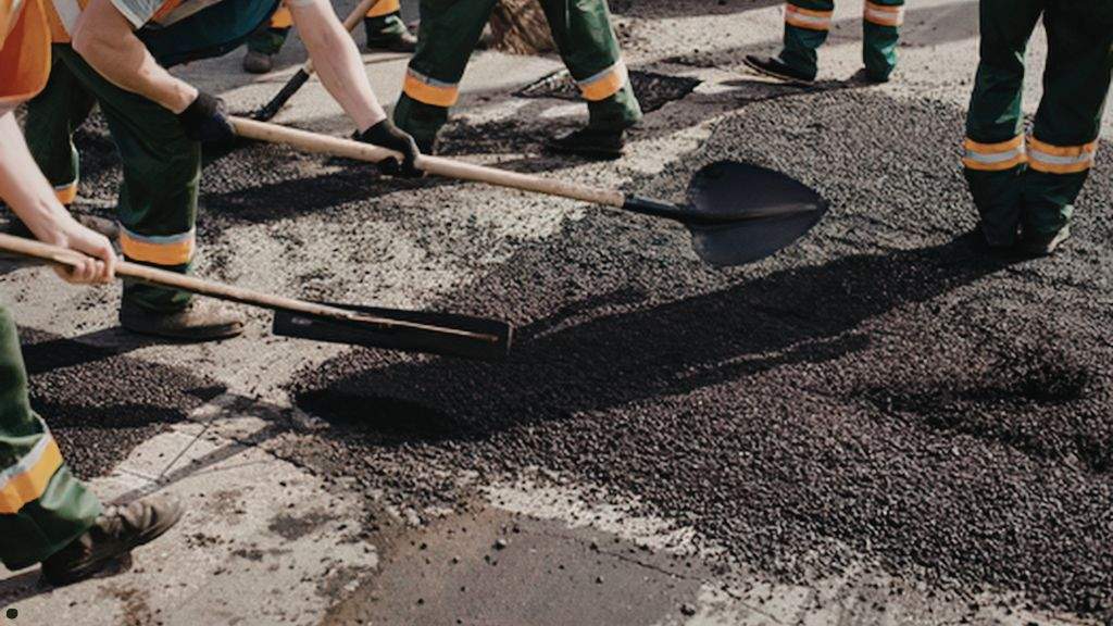 Paving crew spreading and leveling hot-mix asphalt aggregate to patch pavement before fall rain and cooler weather.